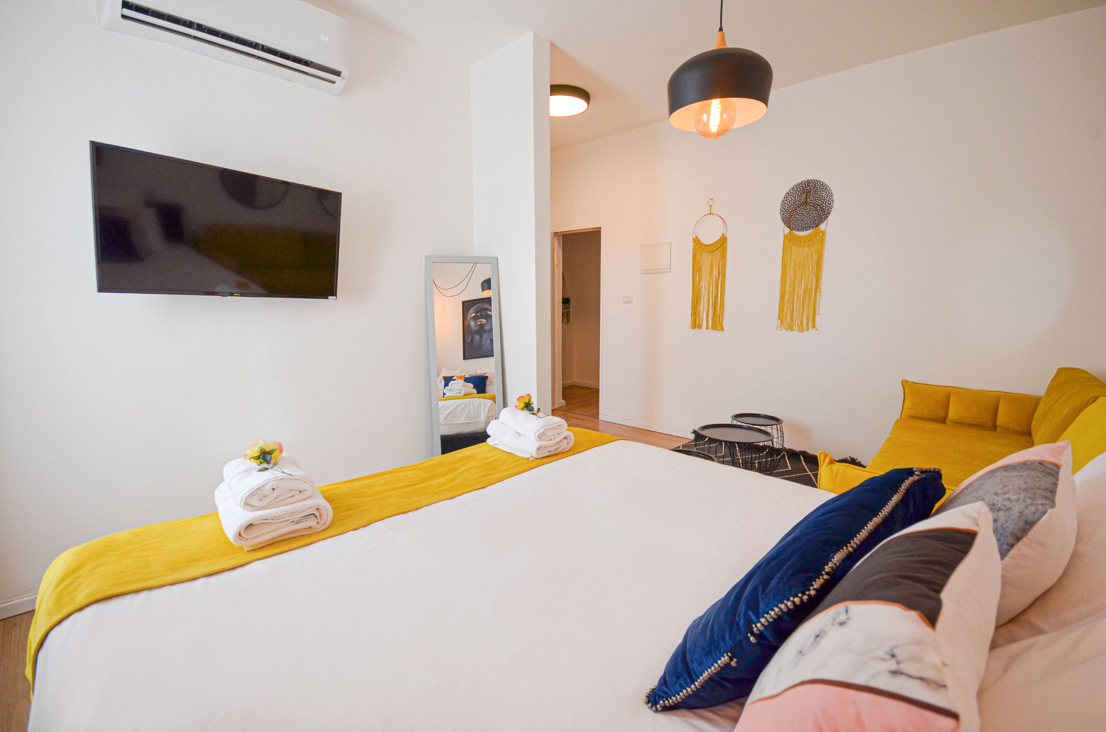 A hotel room with a large bed, a flat-screen TV mounted on the wall, a mirror, and a hanging light fixture.