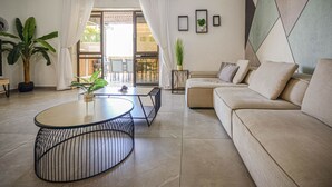 A modern living room with a beige sofa, a round coffee table, and a dining area with chairs. There are potted plants and a view of the outdoors through a glass door. - YalaRent Caesar's Villa - Families only (Eilat)