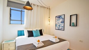 A hotel room with a large bed, a bedside table, a window with curtains, and a wall-mounted picture. - YalaRent Caesar's Villa - Families only (Eilat)