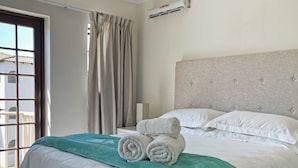 A neatly made bed with white linens and a teal throw blanket, a window with curtains, and an air conditioning unit on the wall.