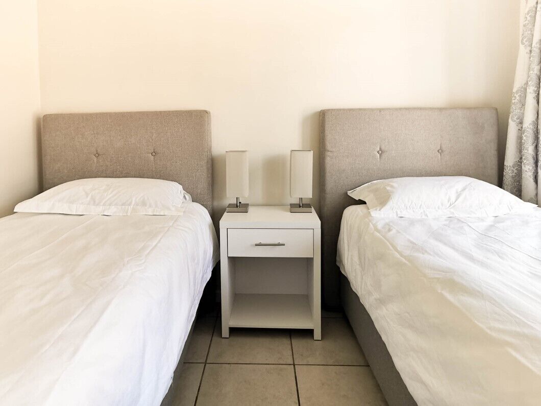 A hotel room with two single beds, each with a white bedspread and matching headboard. There is a nightstand between the beds with a lamp and a drawer.