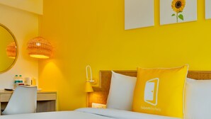 A hotel room with a yellow wall, a bed with white bedding, a bedside table with a lamp, and a chair. There are two framed pictures on the wall, one of a sunflower and one of a sunflower with a leaf. - Bloom Hotel Juhu (Mumbai)