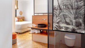 Moderni hotellihuone, jossa on sänky, puinen yöpöytä, jossa on kulho, seinälle kiinnitetty hylly ja suuri kehystetty kuva trooppisesta maisemasta.