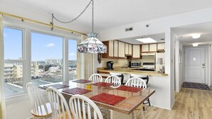 Cozinha moderna com armários brancos, sala de jantar com mesa de vidro e cadeiras brancas, e vista da cidade através de amplas janelas.