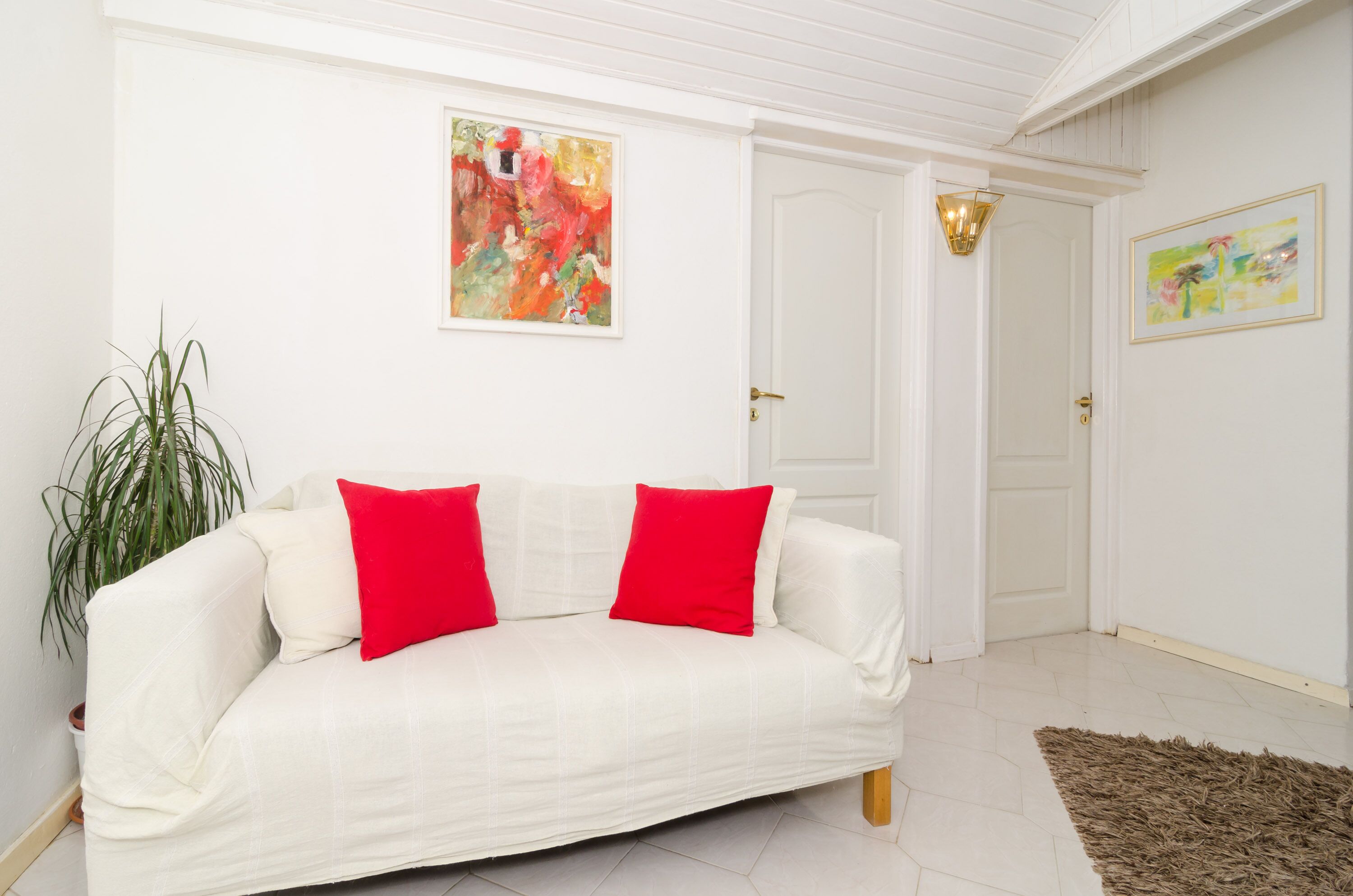 A white sofa with red cushions, a potted plant, and two abstract paintings on the wall.