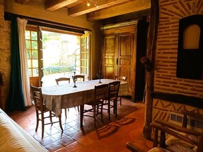 Uma sala de jantar com mesa posta para quatro pessoas, cadeiras de madeira, toalha de mesa estampada e vista para o jardim através de portas abertas.