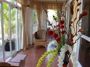 Um quarto com uma cadeira de vime, uma porta de vidro que dá para o exterior e um vaso com flores brancas e vermelhas.