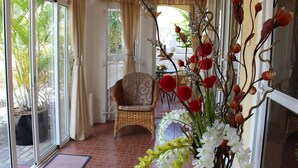 A room with a wicker chair, a glass door leading outside, and a potted plant with white and red flowers.