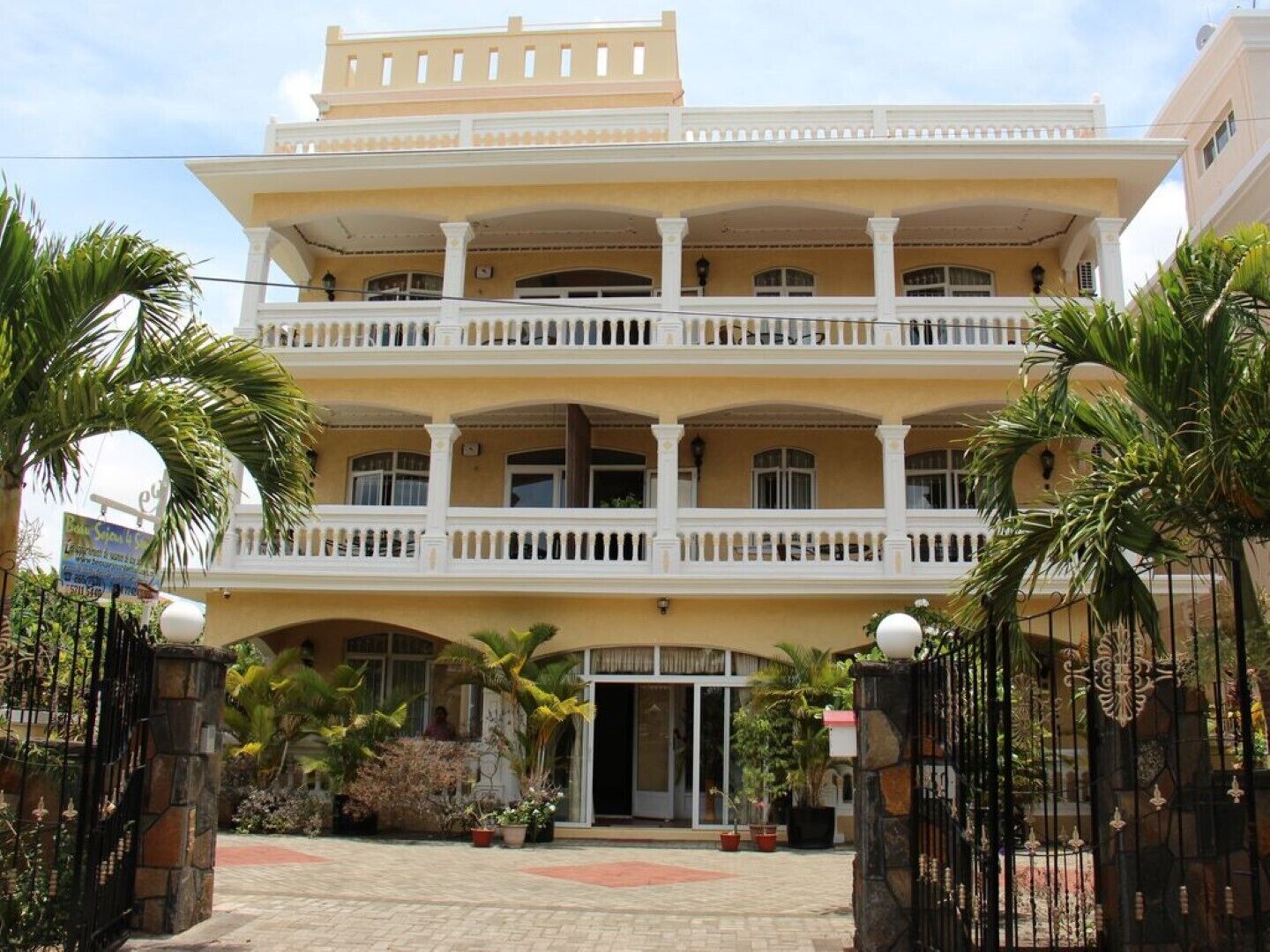 A multi-story hotel with a central entrance, balconies, and a paved courtyard with palm trees.