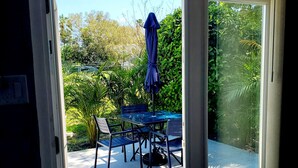 Une porte-fenêtre coulissante donne sur un patio extérieur agrémenté d’une table et de chaises, d’un parasol bleu et d’une végétation luxuriante.