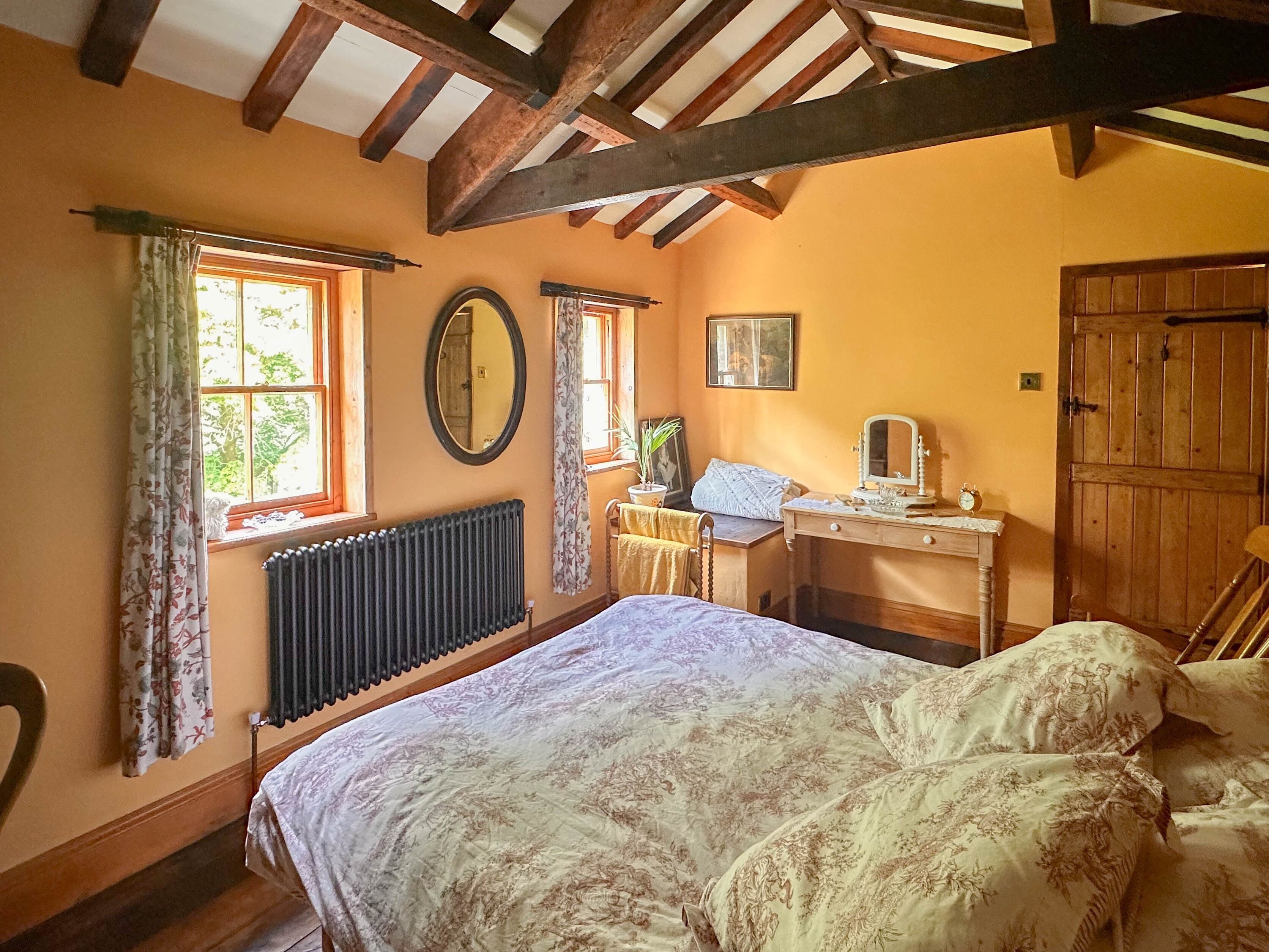 A bedroom with a bed, a desk, a chair, a radiator, a window with curtains, and a wooden door.