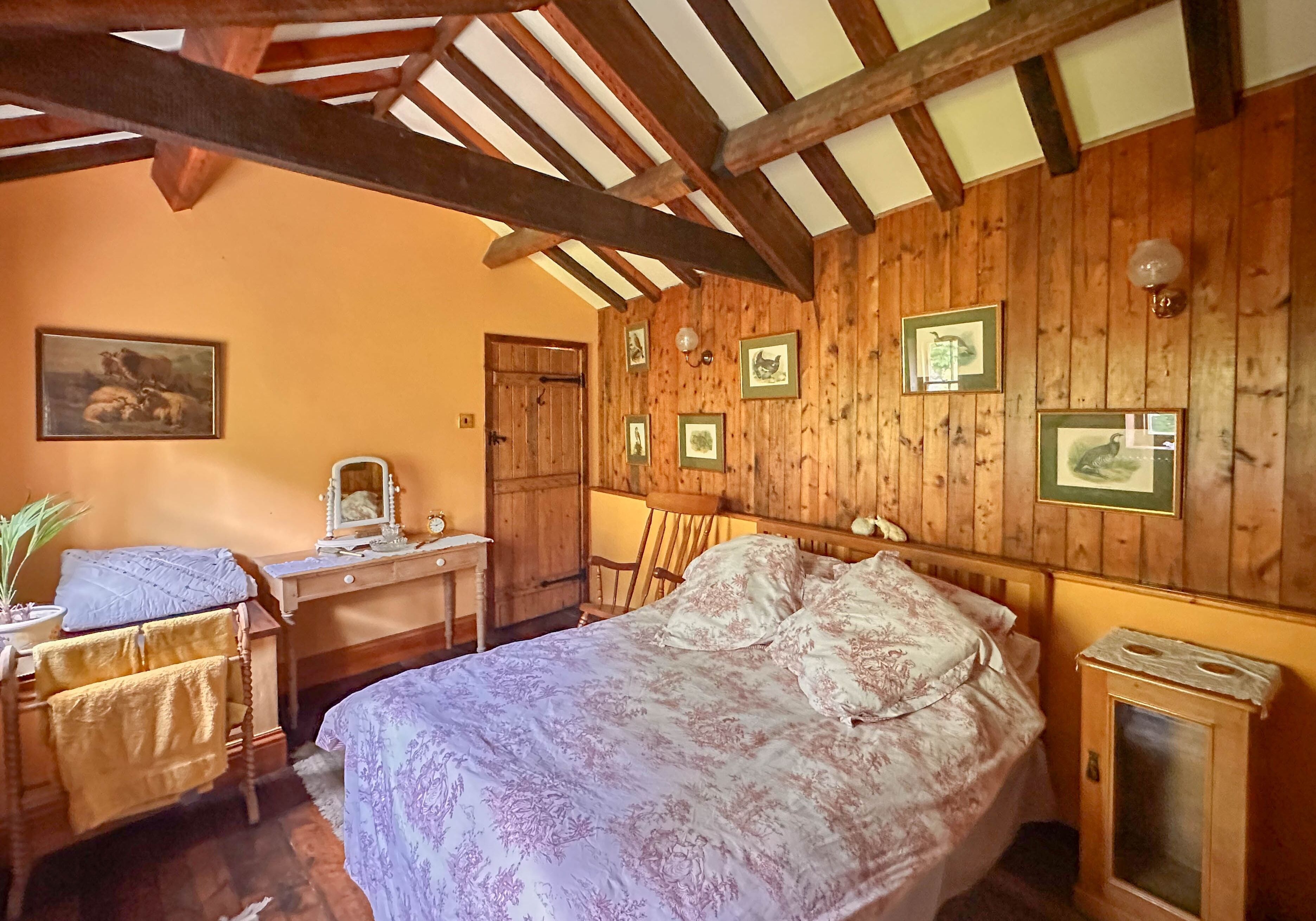 Um quarto acolhedor com teto de madeira, cama com roupa de cama floral, escrivaninha com espelho e quadros nas paredes.