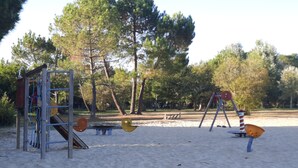 Een speeltuin met schommels en een glijbaan in een zandbak, omringd door bomen.