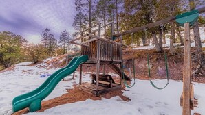 A wooden playground with a slide and swings set in a snowy forest.