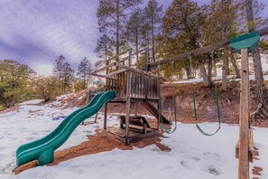 Children can play on the wooden playground structure with a slide and swing set amidst a forested area.