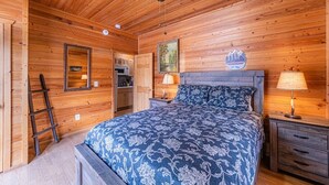 A wooden cabin bedroom with a bed, a nightstand, a lamp, a ladder, and a painting on the wall.