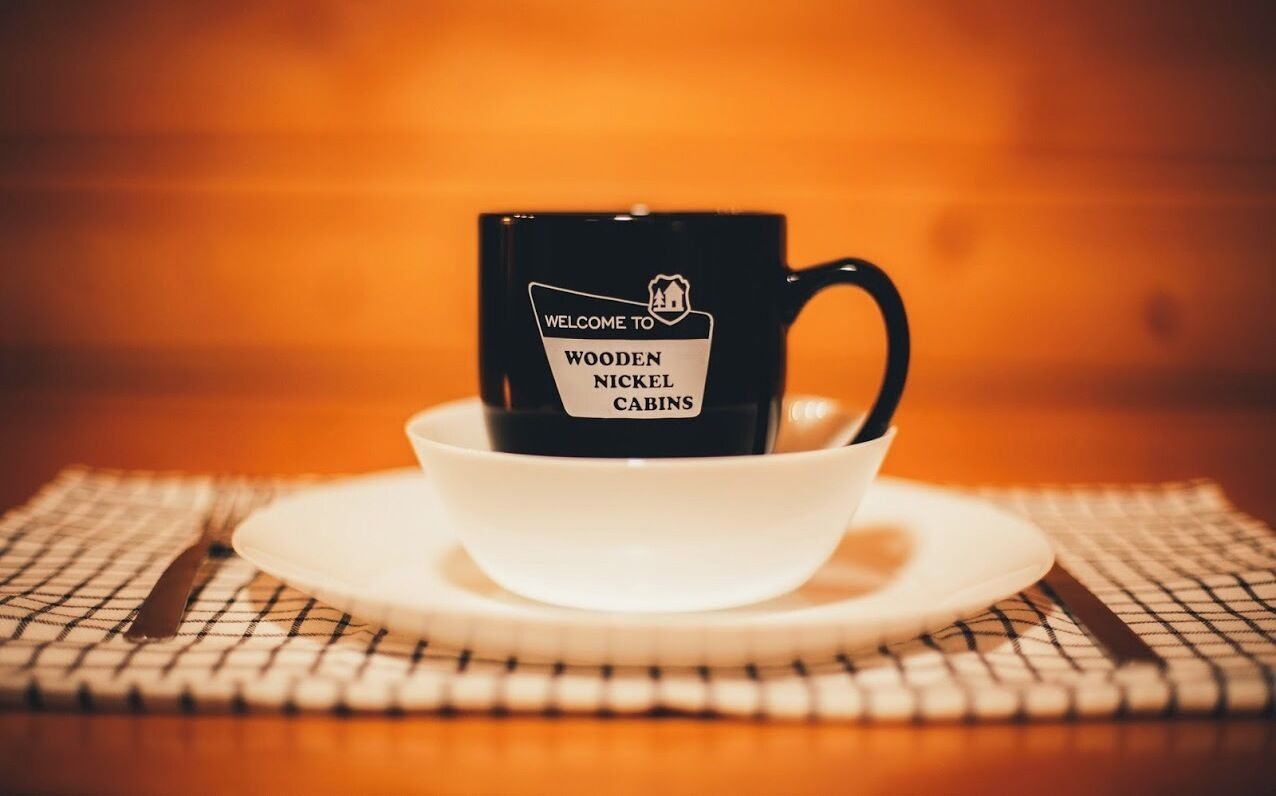 A black mug with "WELCOME TO WOODEN NICKEL CABINS" text, placed on a white saucer and bowl on a checkered placemat.