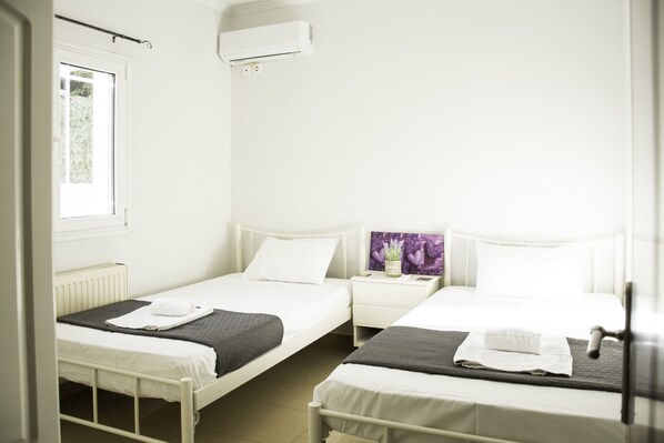 A room with two single beds, a small bedside table with a lamp, and a window with blinds. - Villa Eros (Corfu)