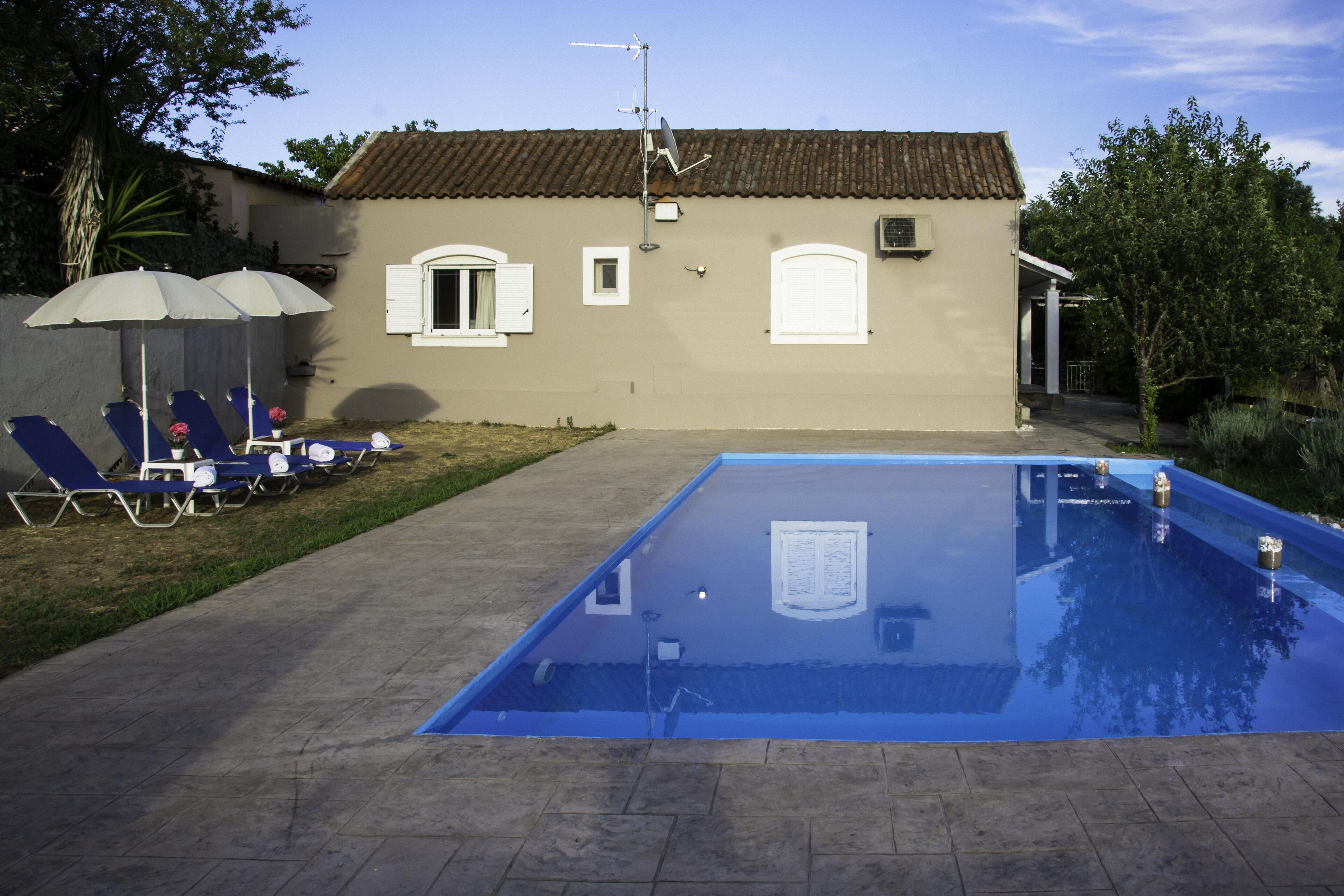 En swimmingpool med solstolar och parasoller, ett hus med ett tegeltak och en satellantenn.
