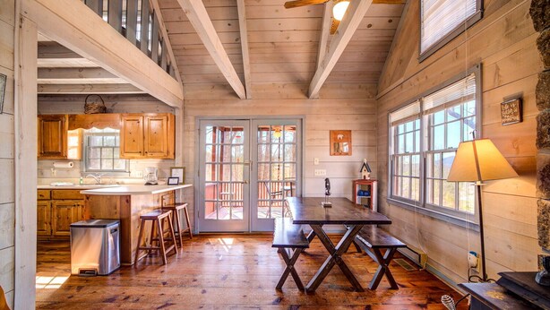 Interior rústico de uma cabana, com uma mesa de jantar de madeira, área de cozinha e janelas grandes.