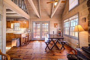 Interno rustico di una baita con un tavolo da pranzo in legno, angolo cottura e ampie finestre.