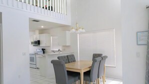 Uma sala de jantar com mesa de madeira e cadeiras estofadas em cinza, cozinha ao fundo e varanda com grades brancas.