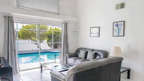 Uma sala de estar com um sofá cinzento, uma mesa de centro de vidro e vista para uma piscina através de amplas janelas.