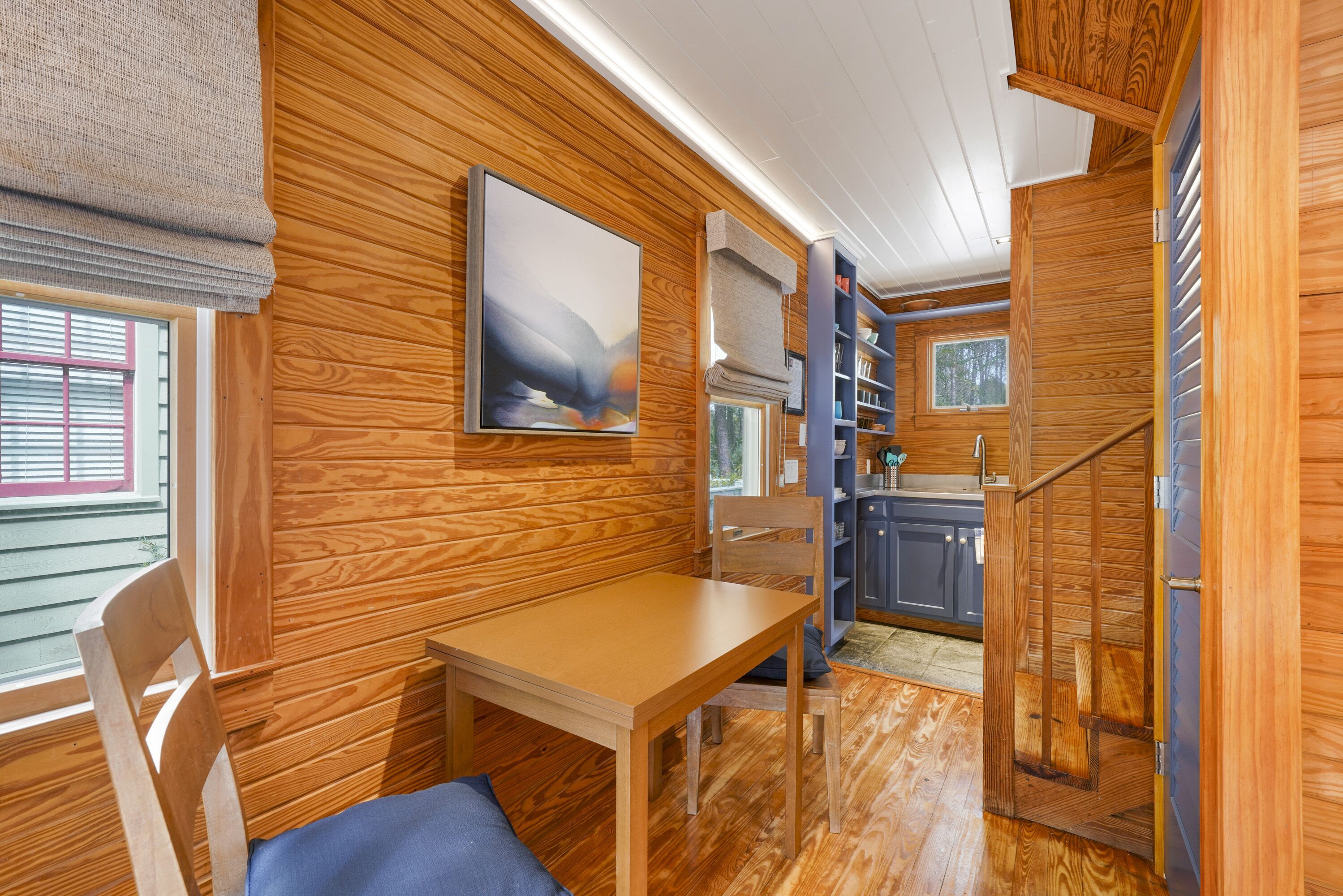A wooden interior with a dining table, chairs, a bookshelf, and a window with blinds.
