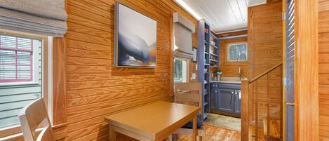 A wooden interior with a dining table, chairs, a bookshelf, and a window with blinds.