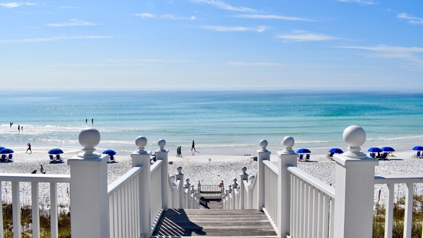 Een strand met een houten looppad dat naar het strand leidt, blauwe parasols en witte leuningen.