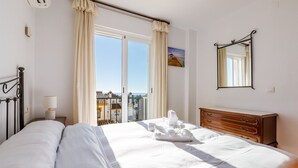 A hotel room with a bed, a wooden dresser, a nightstand with a lamp, a window with curtains, and a painting on the wall. - A&n Moriles 2      . (Torre del Mar)