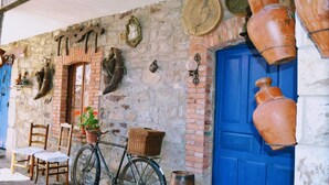 Een landelijk stenen gebouw met een blauwe deur, houten stoelen, een fiets met mandje en diverse aardewerkobjecten aan de muur.