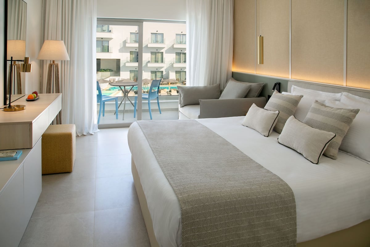 A modern hotel room with a large bed, a desk with a lamp, a wicker ottoman, and a view of the pool and buildings outside.