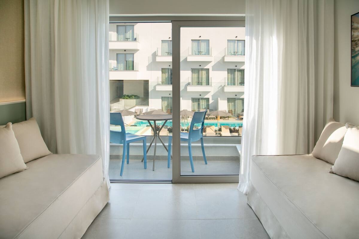 A modern hotel room with a sliding glass door leading to a balcony with a pool and outdoor seating.