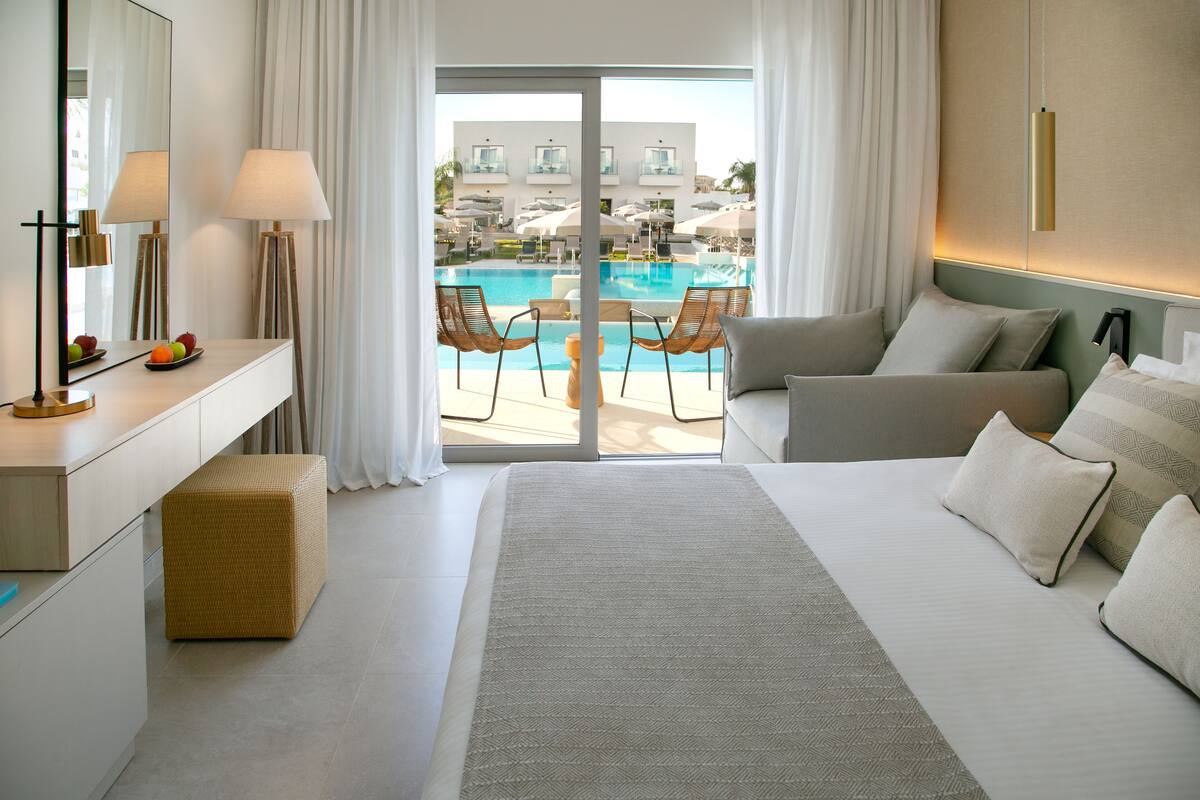 A modern hotel room with a large bed, a bedside table, a mirror, and a view of a pool and outdoor seating area.