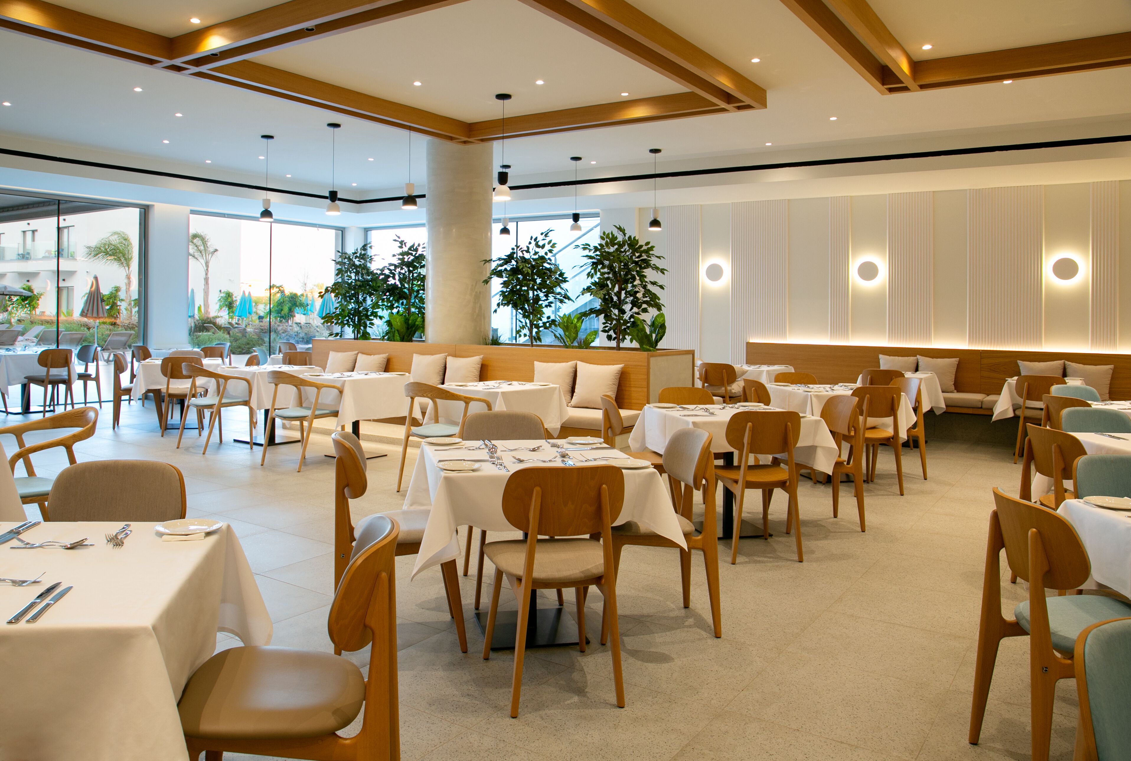 A spacious dining area with multiple tables and chairs, large windows, and indoor plants.