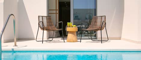 A poolside area with two wicker chairs and a small table with drinks, a pool, and a building entrance.