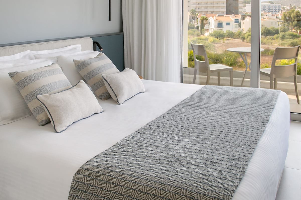 A modern hotel room with a large bed, a balcony with a table and chairs, and a city view.