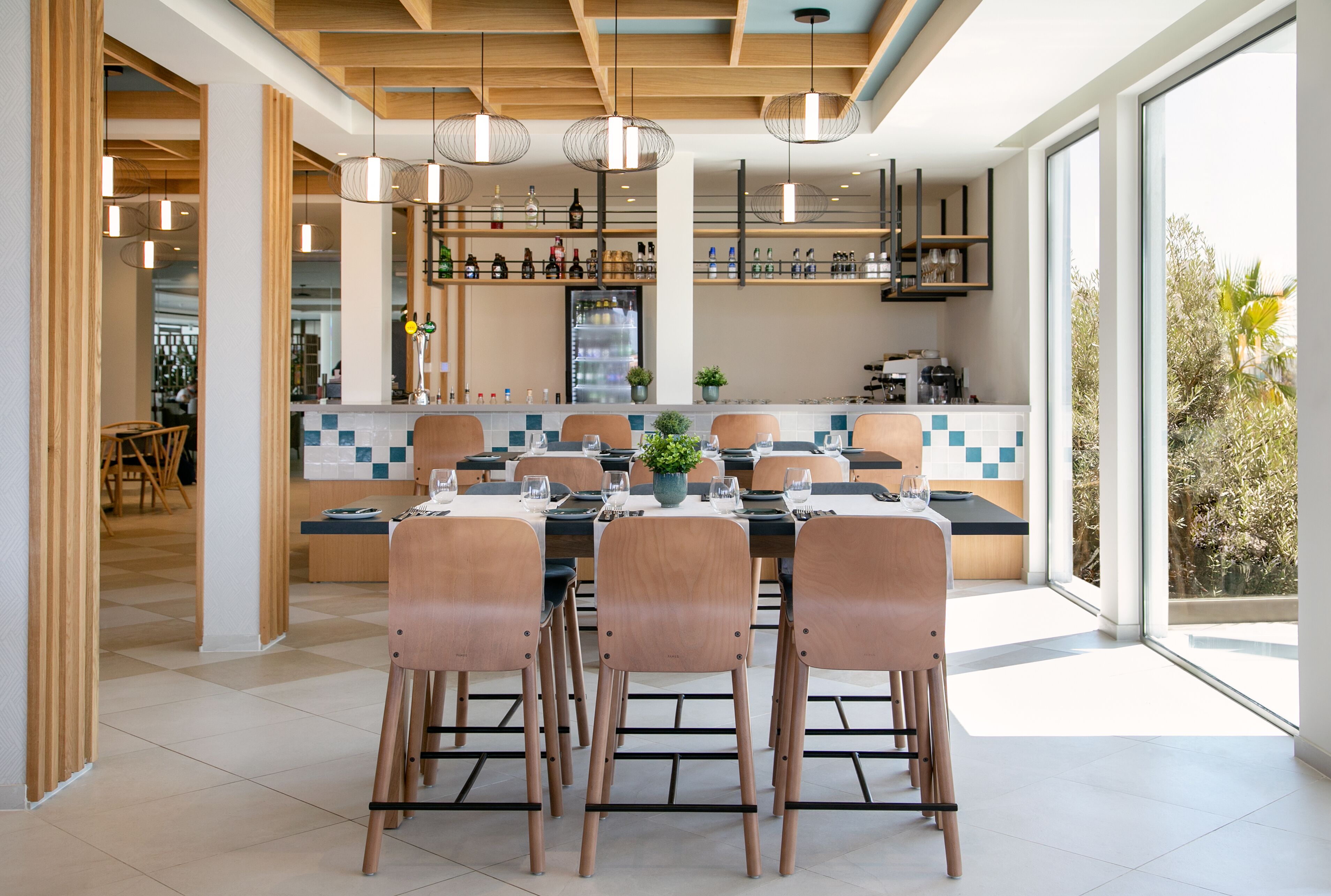 A modern dining area with wooden chairs and tables, a kitchen in the background, and large windows.