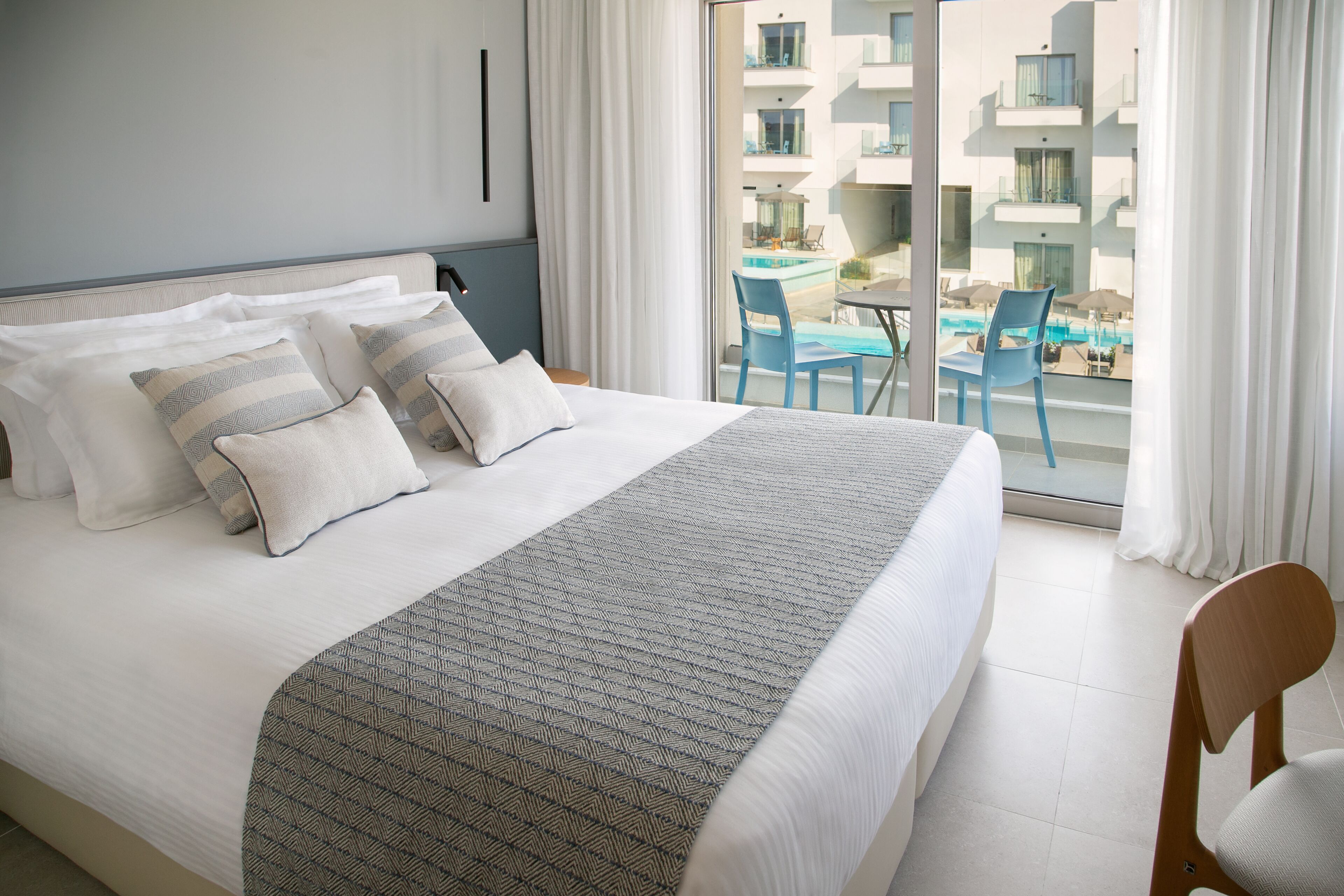 A hotel room with a large bed, a view of the pool and other buildings, and a balcony with chairs.