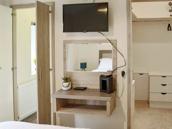 A bedroom with a TV mounted on the wall, a bedside table with a lamp, a small plant, and a bedside console. - Ocean Glade - UK32963 (Corton, near Lowestoft)