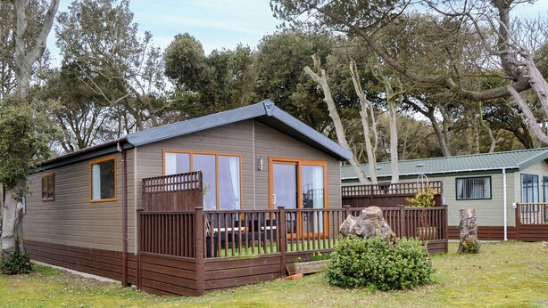 A small, single-story mobile home with a fenced deck, surrounded by trees and grass. - Ocean Glade - UK32963 (Corton, near Lowestoft)