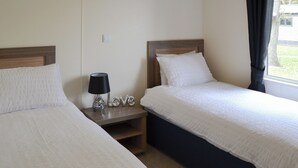A twin room with two beds, a nightstand with a lamp, and a window with curtains. - Ocean Glade - UK32963 (Corton, near Lowestoft)