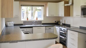 A compact kitchen with a built-in dishwasher, a microwave, and a stove. There is a window with potted plants on the windowsill. - Ocean Glade - UK32963 (Corton, near Lowestoft)