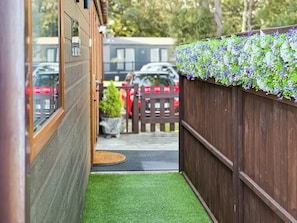 Un étroit sentier menant à une porte en bois, un petit jardin agrémenté de gazon synthétique et une clôture en bois ornée de plantes suspendues.