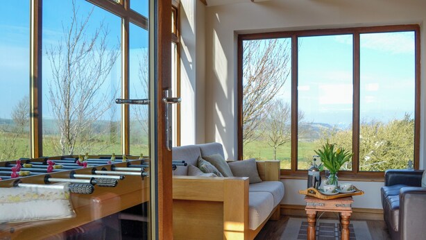 A cozy living room with a sofa, a coffee table, and a foosball table. Large windows offer a view of the outdoors. - 2 bedroom accommodation in Lochmaben near, Lockerbie (Lochmaben near, Lockerbie)