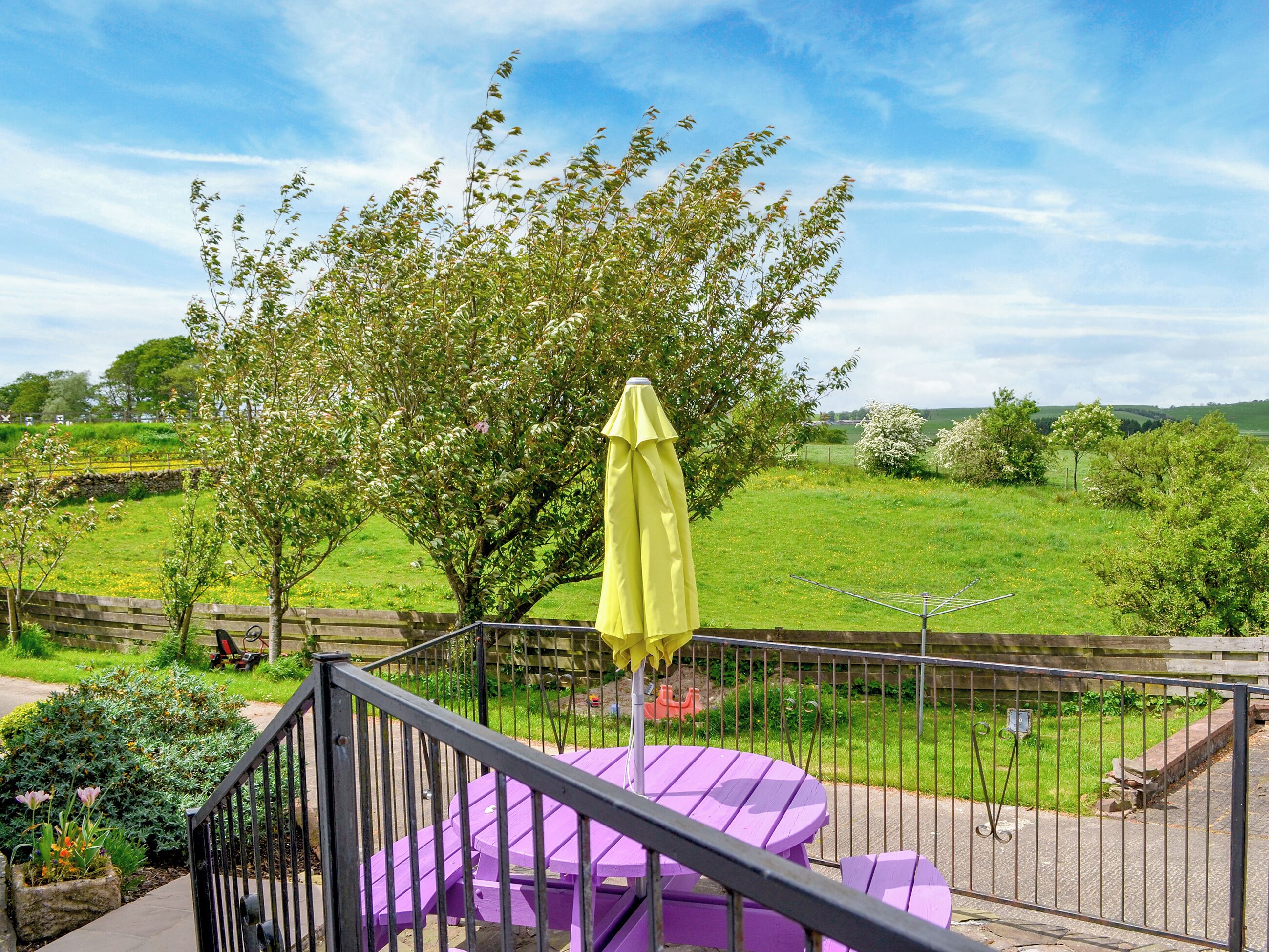 A balcony with a purple table and chairs, a yellow umbrella, and a view of a green field and trees.