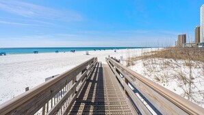 Une promenade en bois mène à une plage de sable fin, bordée d’une eau d’un bleu limpide, avec quelques bâtiments visibles au loin.
