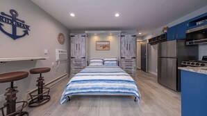 Un dormitorio con cama, escritorio, dos taburetes altos, refrigerador y un reloj en la pared.
