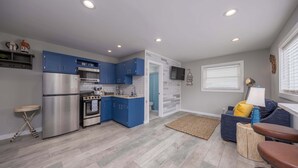 Una cocina compacta con armarios azules, un refrigerador de acero inoxidable y un horno. Hay una pequeña zona de comedor con un sofá azul y un taburete de madera.